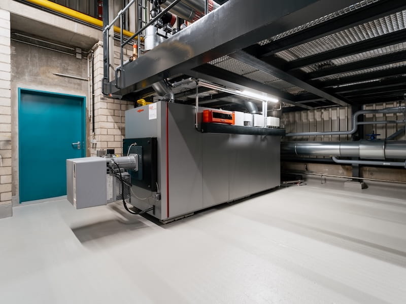 Innenansicht der neu installierten Groß-Wärmepumpe bei der OWS Service für Schienenfahrzeuge GmbH in Weiden. Das Bild zeigt eine moderne Heizungsanlage mit einem großen, grauen Wärmeerzeuger, Rohrleitungen und technischer Infrastruktur. Die saubere und strukturierte Installation befindet sich in einem Technikraum mit Metallgestellen und Zugangstür im Hintergrund.