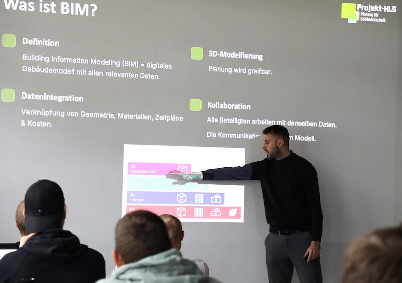 Maximilian Brunner, Prokurist beim Planungsbüro Projekt-HLS aus Weiden, hält an der Dr. Eckert Akademie einen Vortrag über Building Information Modeling (BIM). Er steht vor einer Projektionsfläche, zeigt auf eine Folie mit grafischen Darstellungen und erklärt die Inhalte. Die Präsentation hebt zentrale Aspekte wie Definition, Datenintegration, 3D-Modellierung und Kollaboration hervor. Mehrere Zuhörer sitzen im Publikum und folgen dem Vortrag.