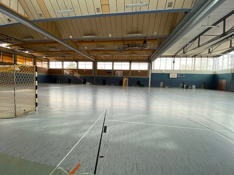 Innenansicht der Turnhalle Eschenbach. Großer, heller Hallenraum mit moderner Deckenbeleuchtung und abgehängter Holzkonstruktion. Sportgeräte wie Handballtor und Basketballkörbe sind sichtbar. Große Fensterfronten sorgen für viel Tageslicht.