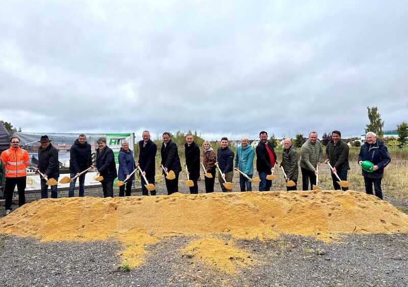 Gruppenfoto beim offiziellen Spatenstich für das neue Designstudio in Selb: Mehrere Personen stehen mit Schaufeln hinter einem Sandhaufen und lächeln in die Kamera. Der Himmel ist bewölkt, im Hintergrund sind Bauzäune und ein Banner zu sehen.