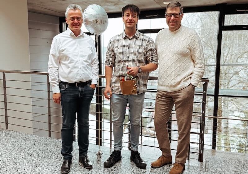 Drei Männer stehen in einem hellen Bürobereich vor einer großen Fensterfront. In der Mitte steht Mitarbeiter Daniel nach seinem erfolgreichen Ausbildungsabschluss zum Technischen Systemplaner für Versorgungstechnik bei Projekt-HLS und hält ein kleines Geschenk in der Hand. Links und rechts neben ihm stehen die Geschäftsführer Jörg Retzer und Jürgen Brunner und lächeln in die Kamera.